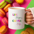 Load image into Gallery viewer, Life Happens Coffee Helps Coffee Mug surrounded by colorful macarons on a bright background.