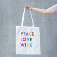 Load image into Gallery viewer, Peace Love Wine Tote Bag featuring colorful lettering on a white tote being held against a gray background.