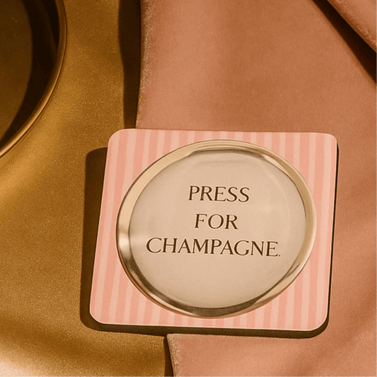 Press for champagne drink coaster on a table, featuring a humorous design with a pink striped border.