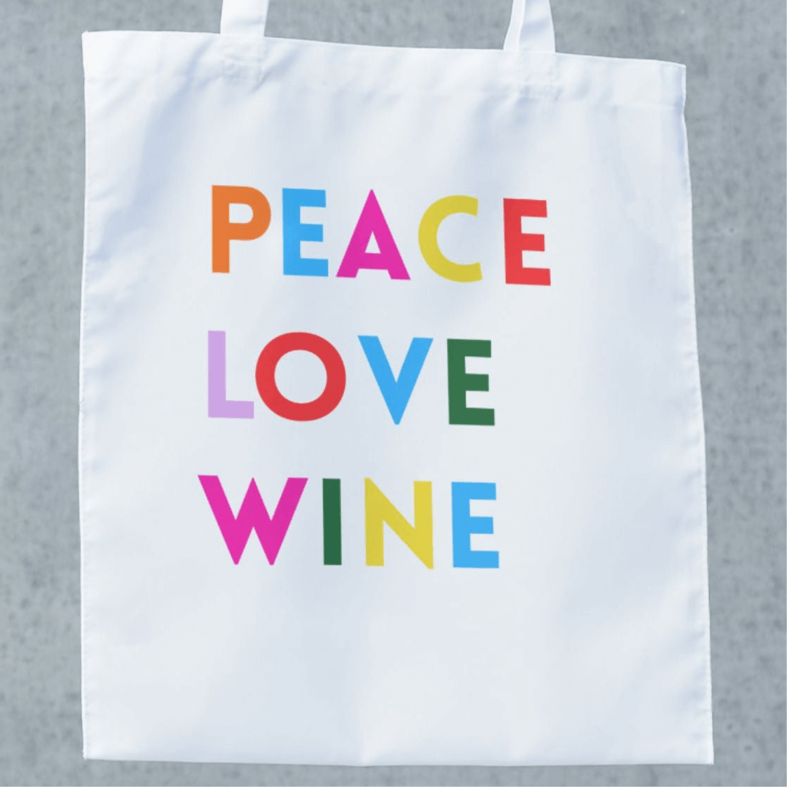 White tote bag featuring colorful text: PEACE LOVE WINE, perfect for wine lovers and trips to the winery.