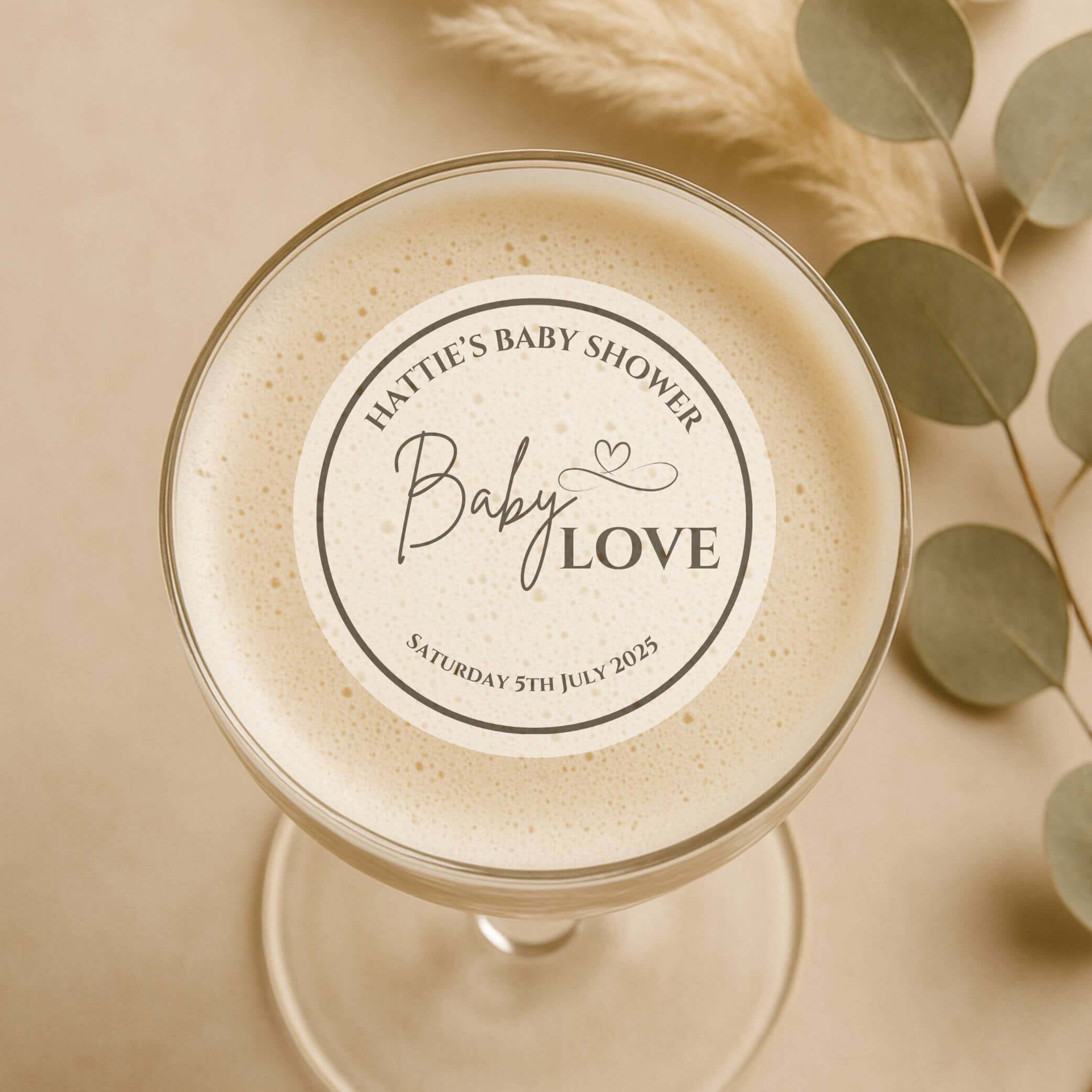Glass with a 'Baby Love' label for a baby shower on a beige background with eucalyptus leaves.