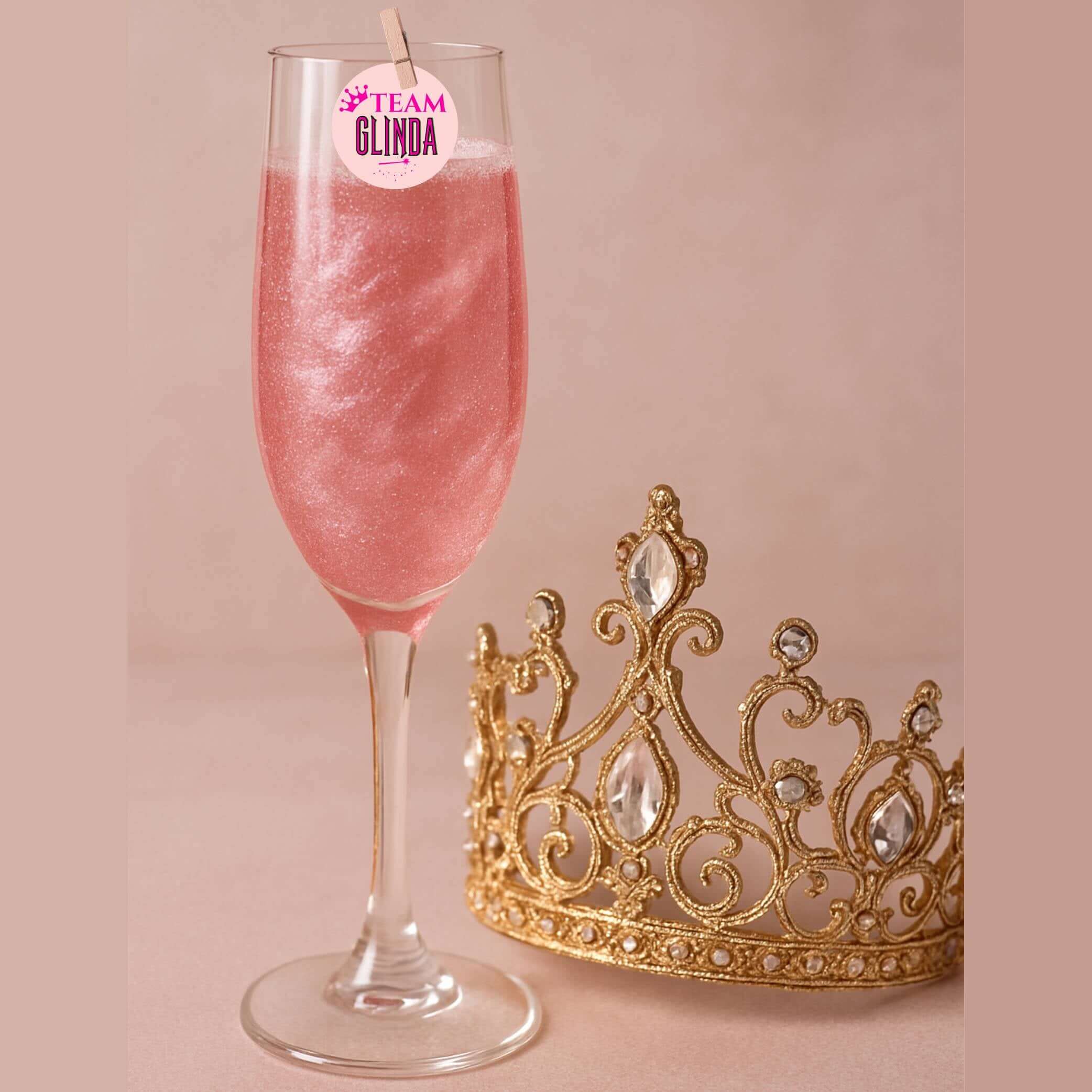 Pink sparkling drink topped with Team Glinda edible topper next to a golden crown.