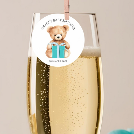 Champagne flute with a baby shower label featuring a teddy bear and gift box on a beige background.