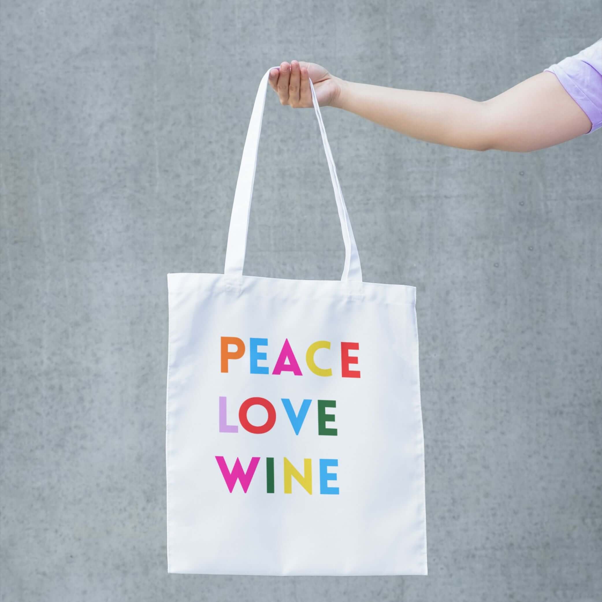 Peace Love Wine Tote Bag featuring colorful lettering on a white tote being held against a gray background.