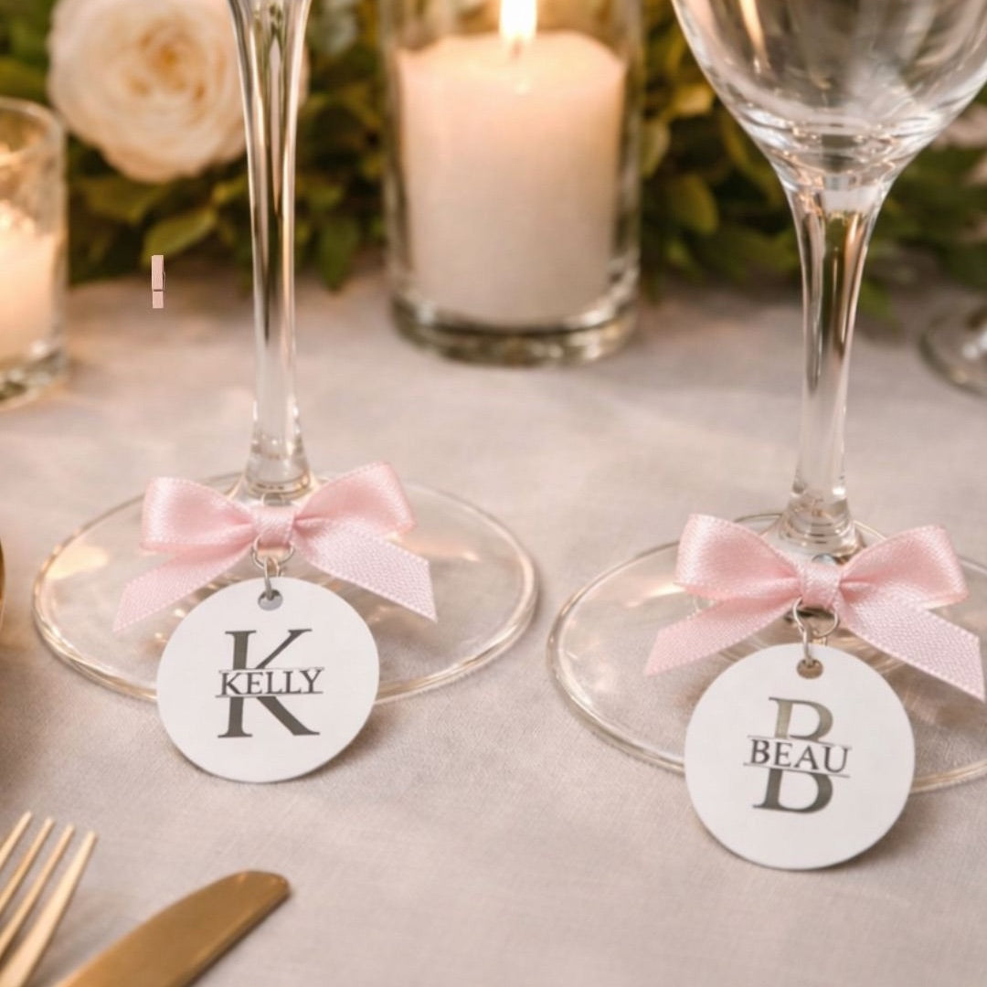 Two wine glasses with decorative tags and pink ribbons on a table setting with candles and cutlery.